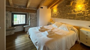 Een slaapkamer met een stenen muur, houten plafondbalken, een raam, een bed met witte beddengoed en een nachtkastje.
