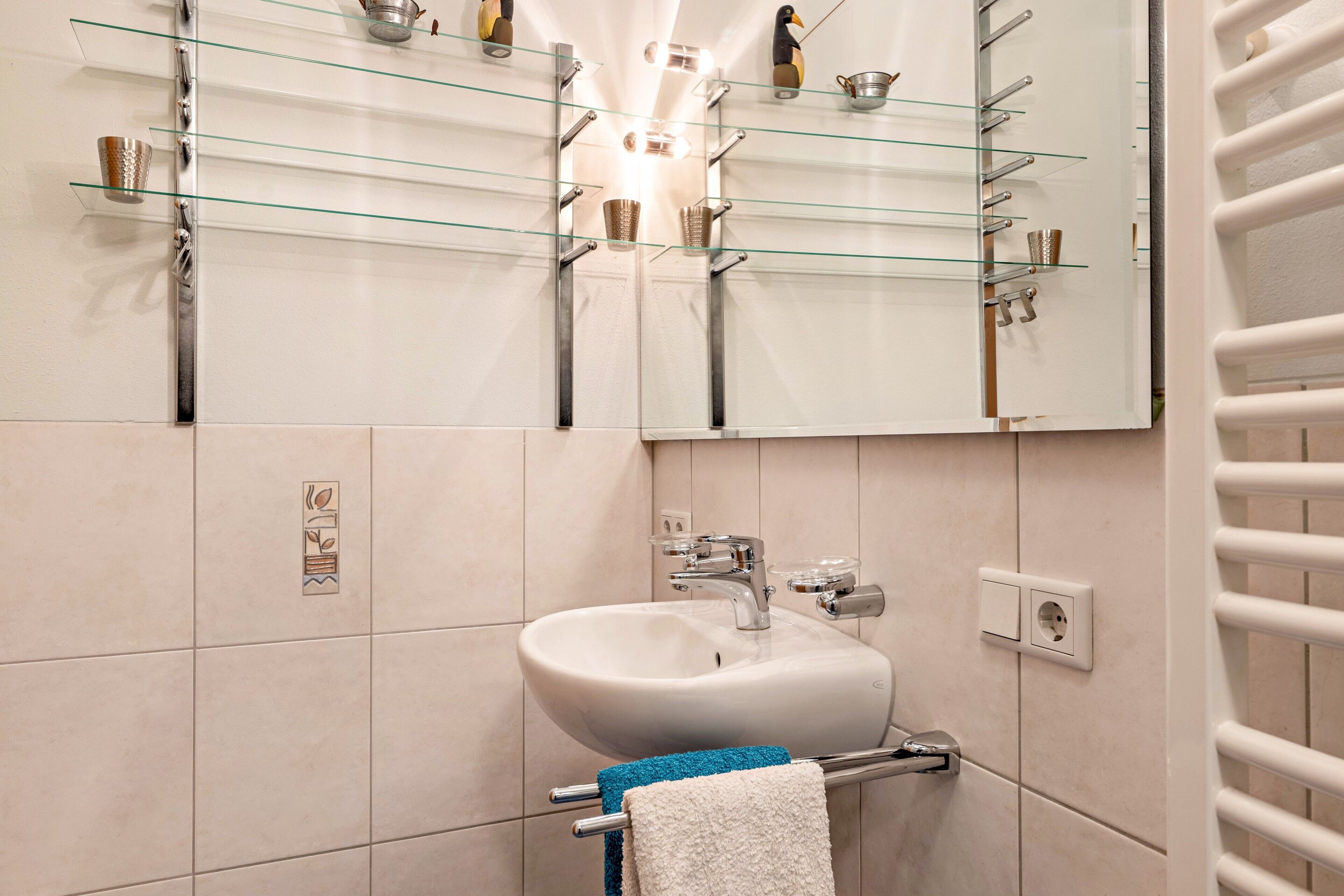 Une salle de bain avec un lavabo blanc, une étagère en verre au-dessus et un porte-serviettes avec des serviettes.