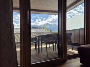 Une pièce dotée d’une porte-fenêtre coulissante donnant sur une terrasse extérieure avec une table et des chaises, une vue sur les montagnes et un plafond en bois.