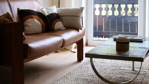 Ein Wohnzimmer mit braunem Ledersofa, einer Stehlampe, einem Couchtisch und Blick durch eine Glastür auf einen Außenbalkon.