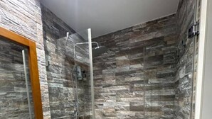 Een badkamer met een wand met steenstructuur, een douche met glazen wanden en een wit wastafelmeubel met houten details.