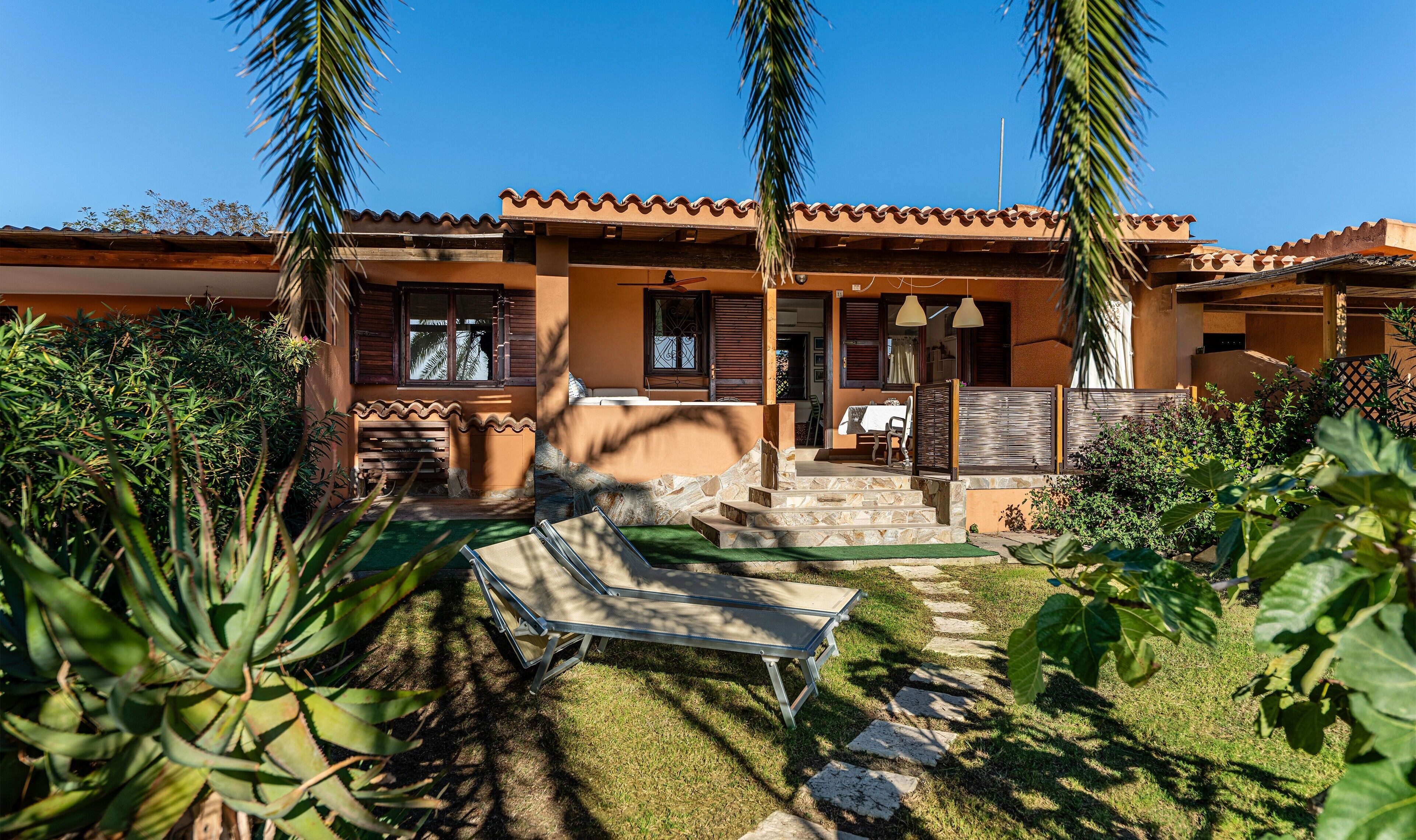 A two-story building with a tiled roof, a patio with a table and chairs, and a small garden area with plants and a sun lounger.