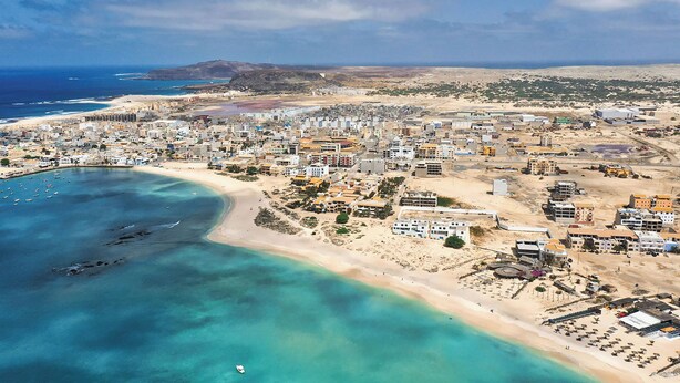 Vista aérea de uma cidade costeira com praia, edifícios e águas azuis cristalinas.
