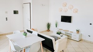 Quarto moderno e bem iluminado, com mesa de jantar, cadeiras, sofá, televisão na parede e um pequeno vaso de planta.