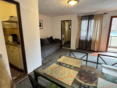 A living room with a glass table, a sofa, a coffee machine, and a view of a balcony.