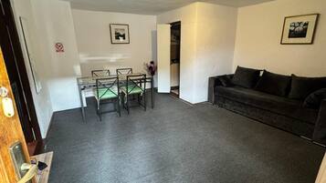 Un salon avec un canapé foncé, une table à manger avec des chaises, et un coin cuisine visible à travers une porte ouverte.