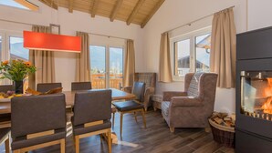 Una habitación acogedora con techo de madera, una mesa de comedor con sillas, un sofá, una chimenea y ventanas grandes con cortinas.