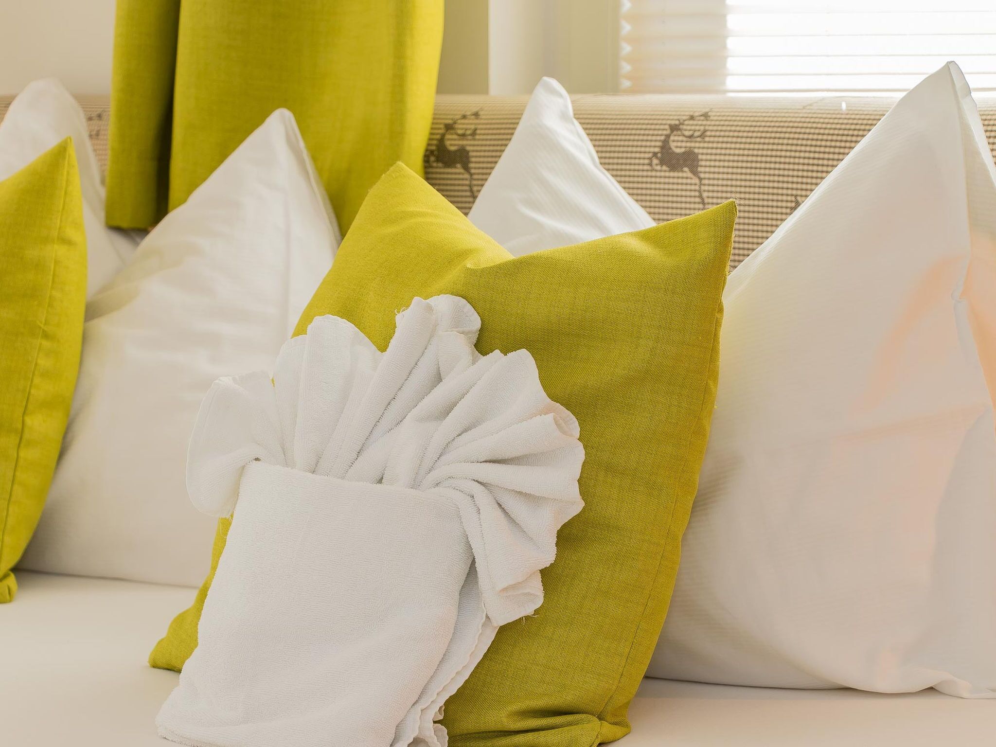 Una cama con almohadas en tonos verde y blanco, una toalla blanca y una ventana con persianas al fondo.