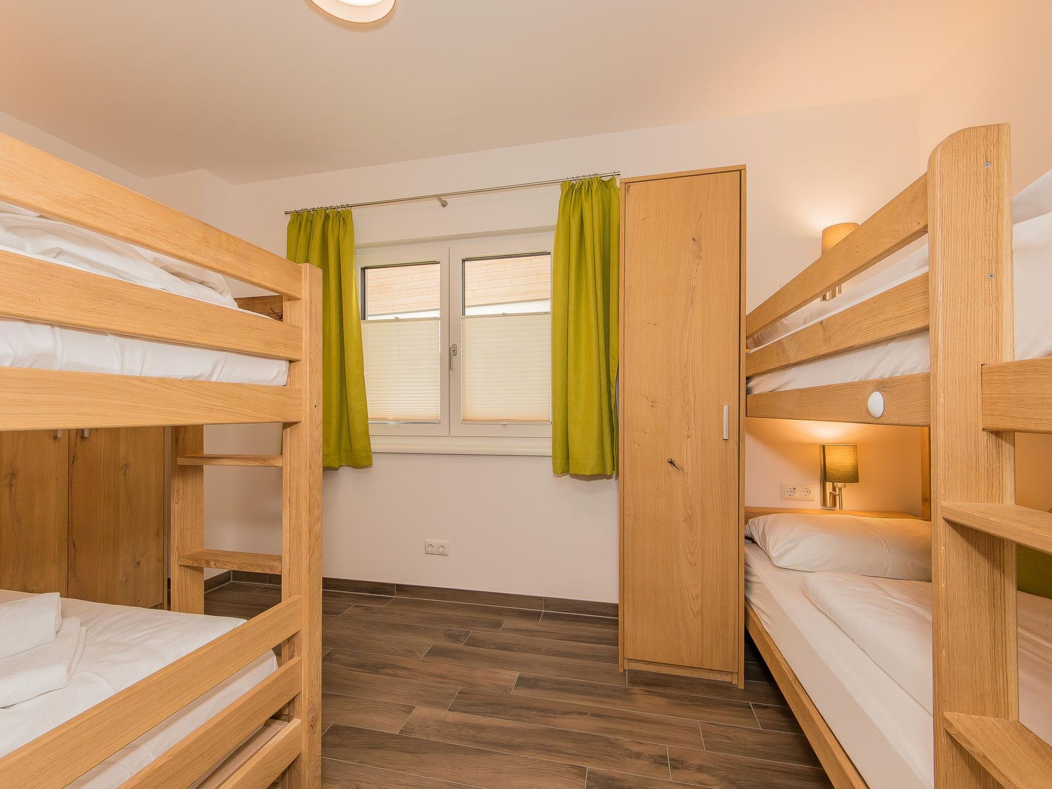 A room with bunk beds, a window with curtains, a bedside lamp, and a wooden wardrobe.