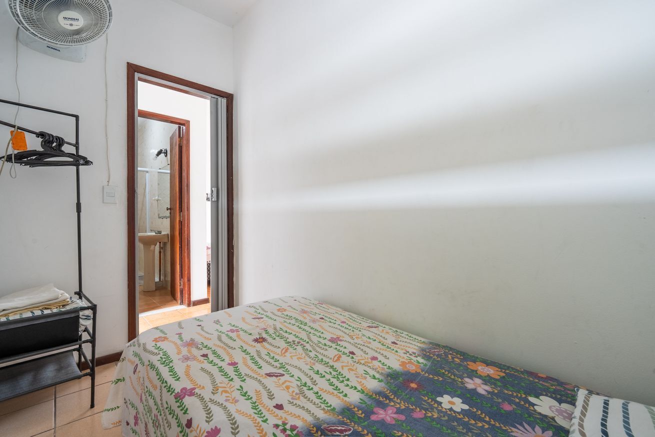 Un dormitorio con cama, un ventilador de pie, una estantería con objetos y una puerta que da al baño.