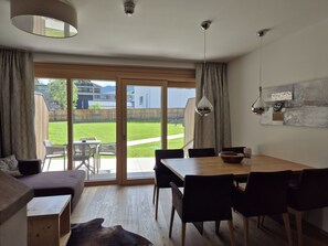 A modern living room with a dining area, large sliding glass doors to a backyard, and a view of neighboring buildings. - 3_SPA - Deluxe Garden 417 by Interhome (Zell am See)