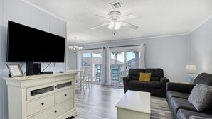A living room with a flat-screen TV, a white cabinet, a dark sofa, a small coffee table, and a ceiling fan. - Floral Villa D14 - Three Bedroom Two Bath Condo with Shared Pool (Surfside Beach)