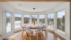 A dining area with a round table, six chairs, and large windows offering a view of a snowy landscape. - Waterfront with Spa - Pool Table - Le Rivage Alpin (Saint-Côme)
