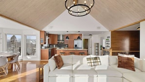 A modern living room with a white sectional sofa, wooden flooring, a large chandelier, and a view of the kitchen and dining area. - Waterfront with Spa - Pool Table - Le Rivage Alpin (Saint-Côme)