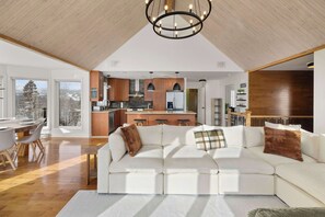 Ein modernes Wohnzimmer mit einem weißen Ecksofa, Holzboden, einem großen Kronleuchter und Blick auf die Küche und den Essbereich.