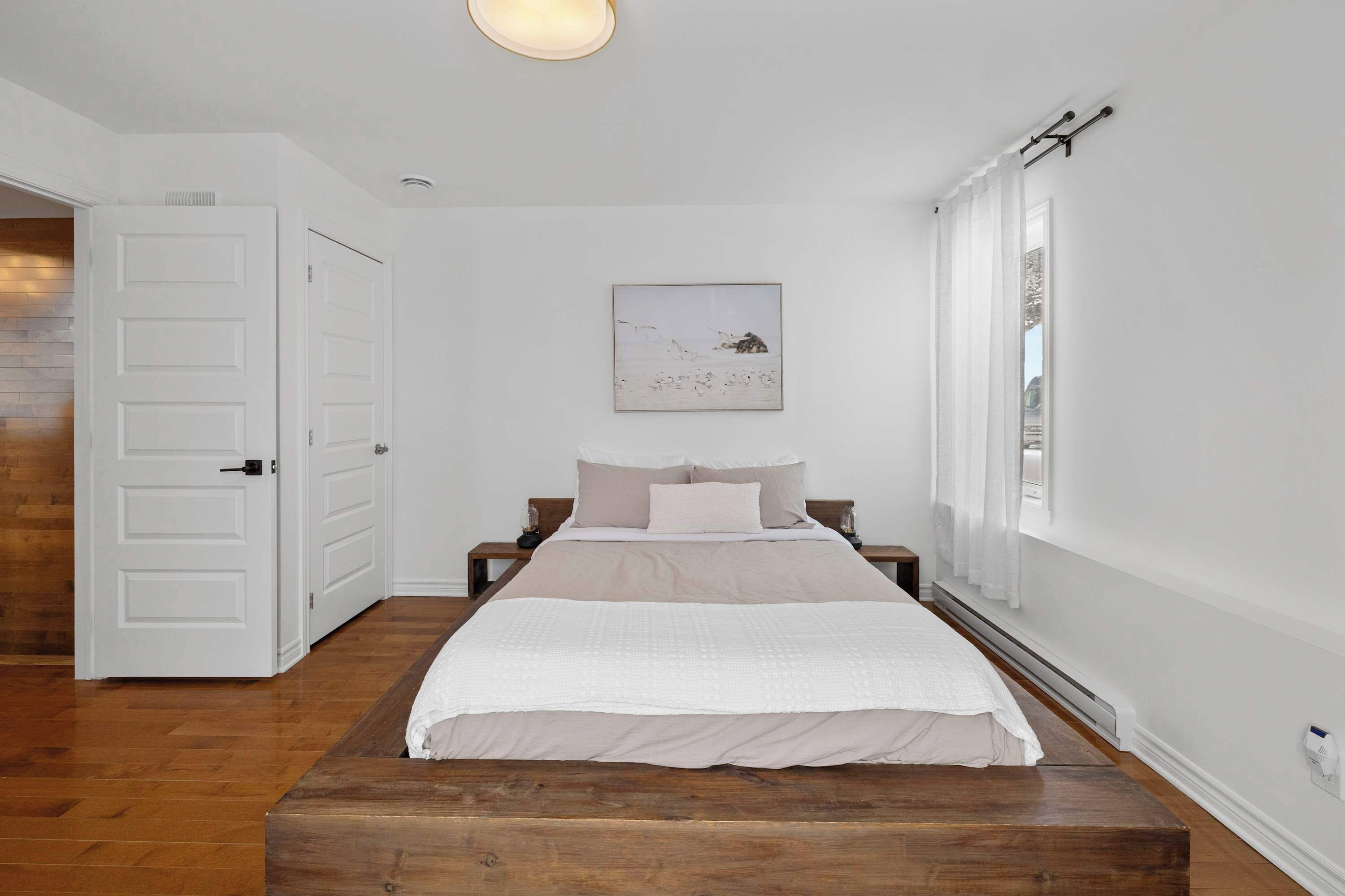 A bedroom with a wooden bed, white walls, a painting, and a window with white curtains.