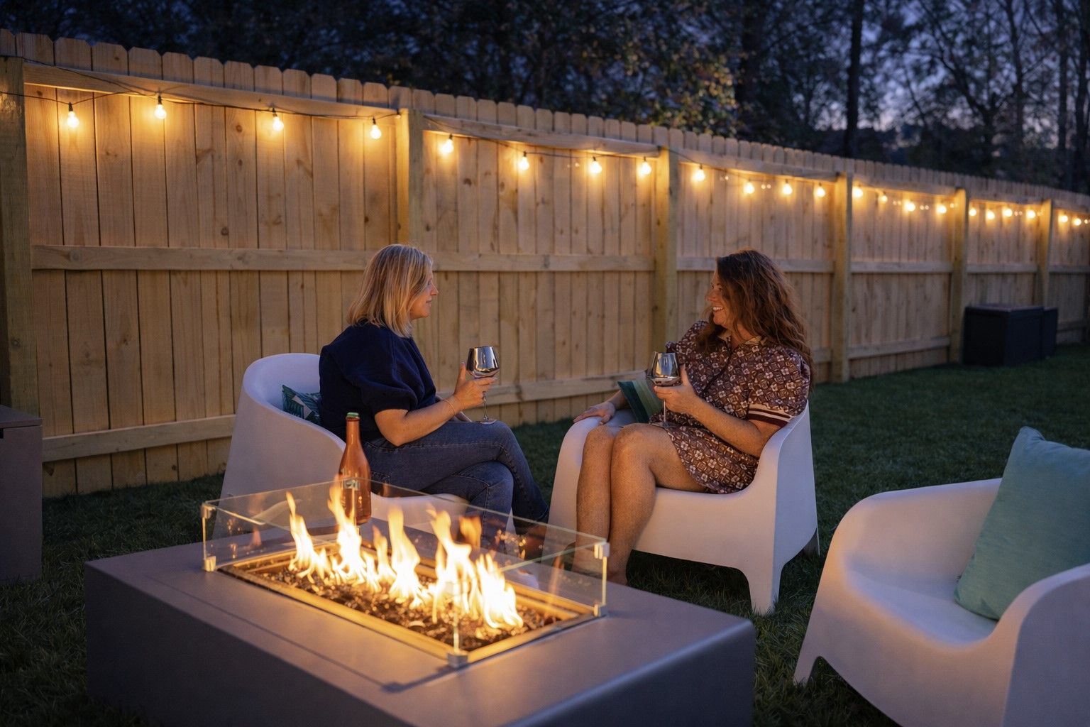 Deux personnes sont assises sur des chaises blanches, face à une table en verre, avec un foyer au centre, le tout éclairé par des guirlandes lumineuses, et une clôture en bois en arrière-plan.