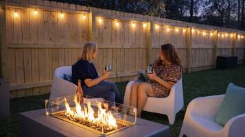 Deux personnes sont assises sur des chaises blanches, face à une table en verre, avec un foyer au centre, le tout éclairé par des guirlandes lumineuses, et une clôture en bois en arrière-plan.