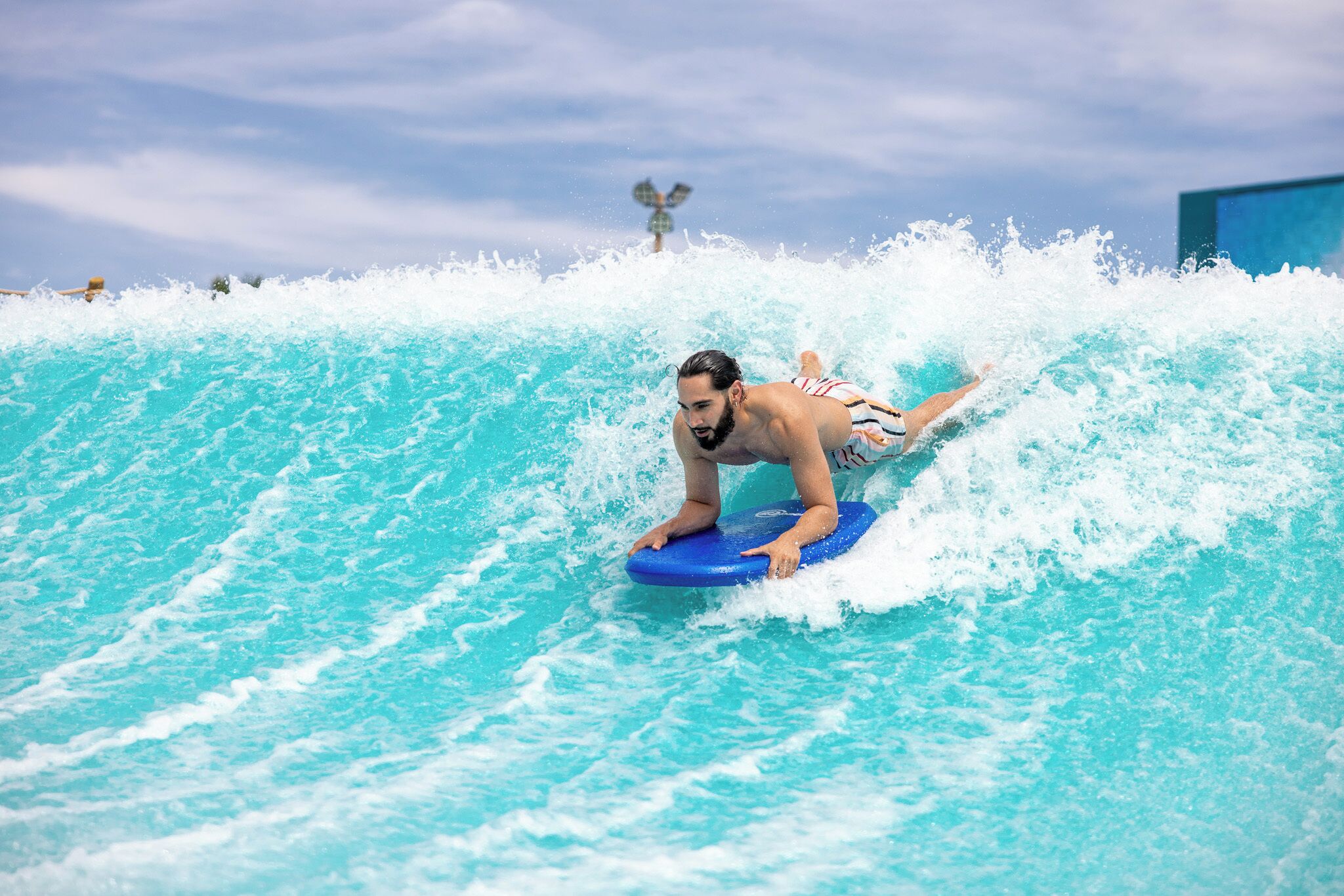 Une personne surfe sur une vague à l’aide d’une planche de surf bleue dans une piscine à vagues.