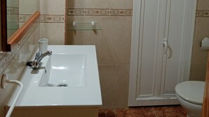 Ein Badezimmer mit weißem Waschbecken, Spiegel und Toilette. Über dem Waschbecken befinden sich Glastage und rechts eine weiße Tür.