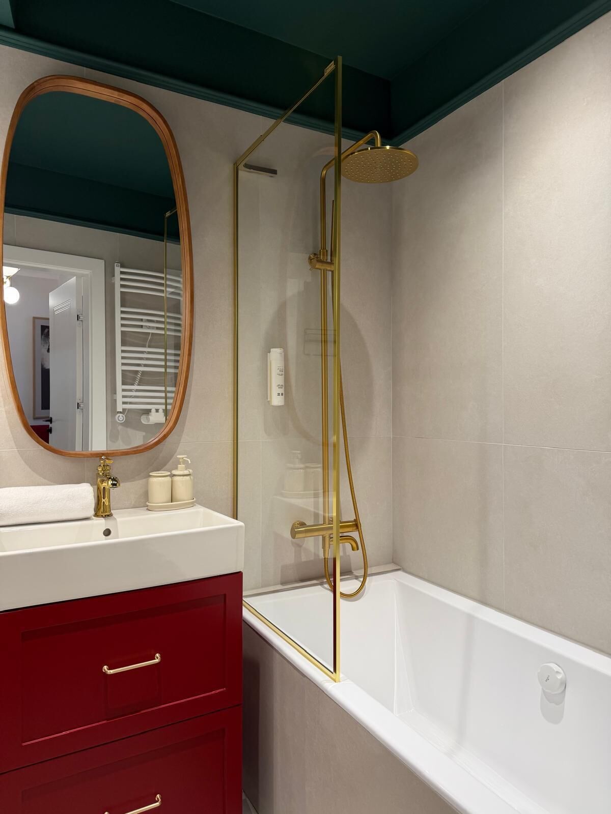 A modern bathroom with a white bathtub, a gold showerhead, a large mirror, and a red vanity with drawers.