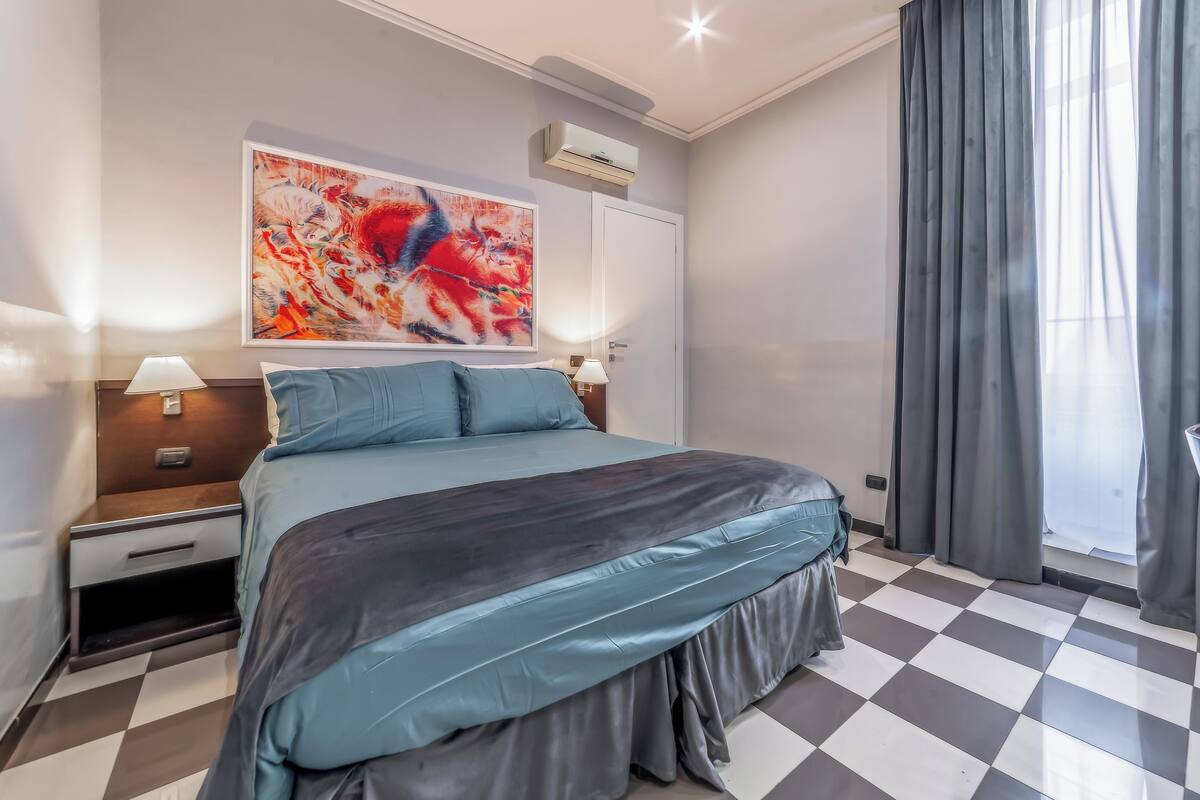 Habitación de hotel con cama, mesita de noche, lámpara, aire acondicionado, ventana con cortinas y un cuadro abstracto grande en la pared.