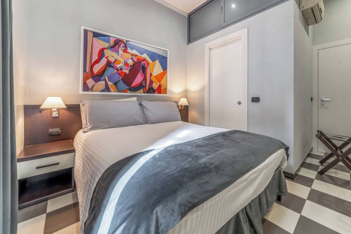 Una habitación de hotel moderna con una cama grande, dos mesitas de noche con lámparas, un cuadro en la pared y un suelo a cuadros.