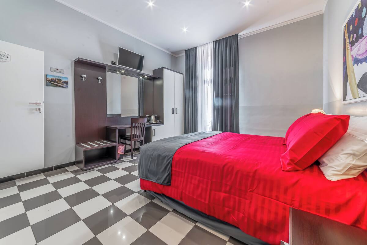 Habitación de hotel con suelo a cuadros, una cama con ropa de cama roja y gris, un escritorio con una silla, un armario y un amplio ventanal con cortinas.