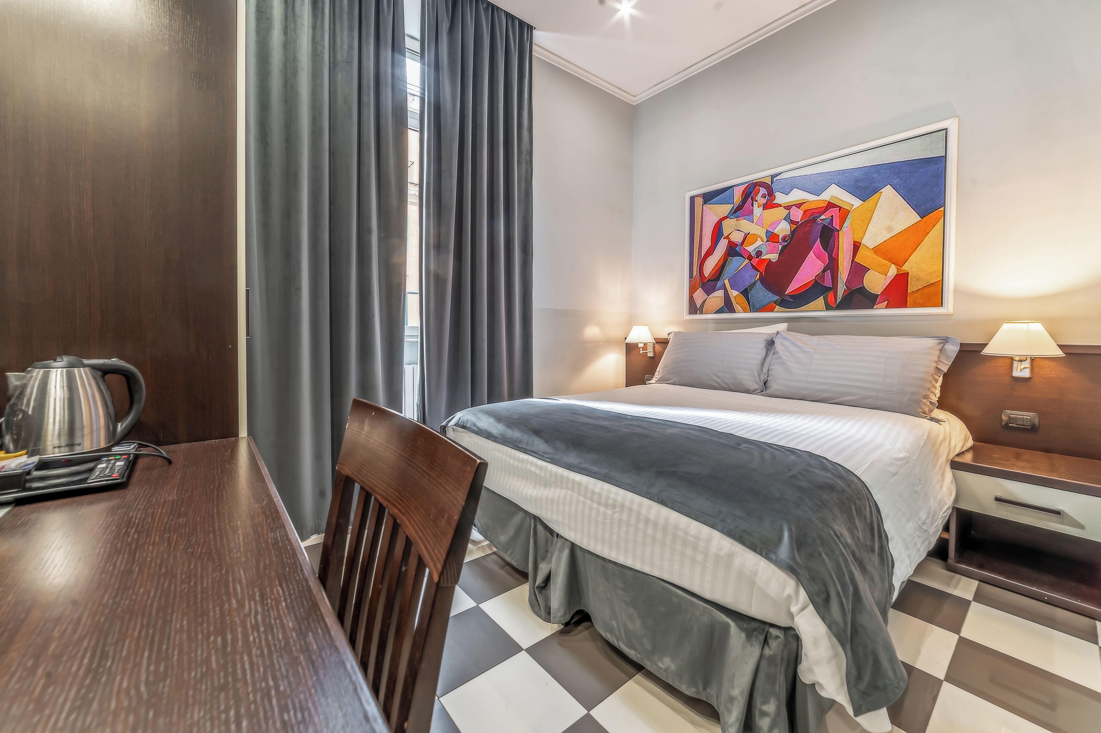 Une chambre d’hôtel avec un lit, un bureau doté d’une bouilloire, une chaise, un tableau au mur et un sol à carreaux.