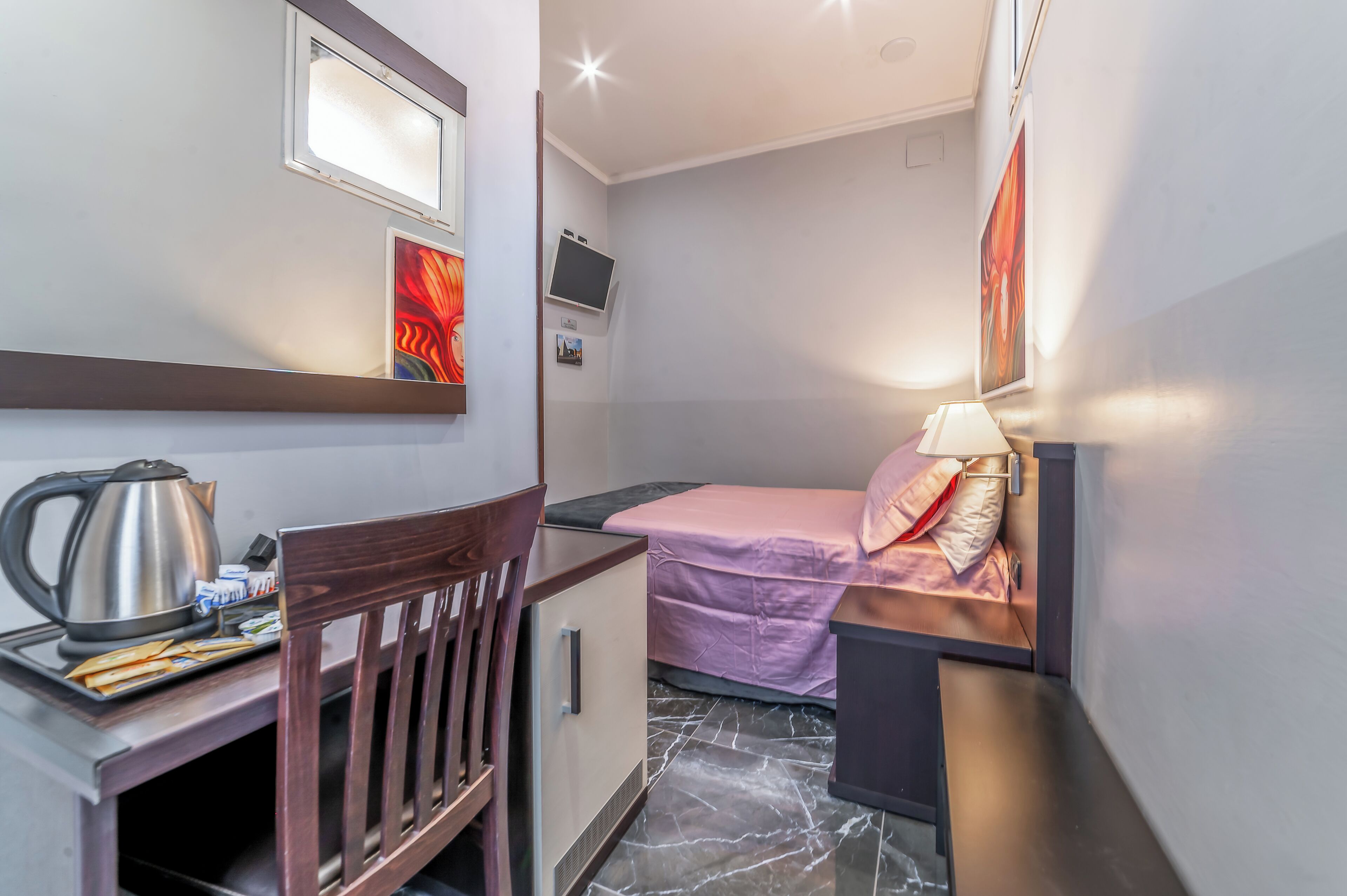 Habitación de hotel compacta con cama, escritorio y silla, un mueble pequeño, televisor y estante con hervidor y algunos objetos.