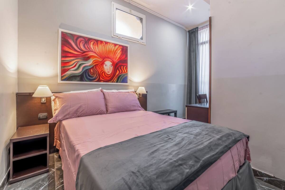 Un cuarto de hotel con cama, dos lámparas de noche, una mesita de noche, una pequeña mesa, un cuadro en la pared y una ventana con cortinas.