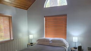 A bedroom with a bed, two bedside tables, a window with blinds, and a wooden ceiling.