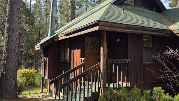 A rustic cabin with a green roof, a chimney, and a wooden porch with a staircase leading up to the entrance. - Cozy Studio in Idyllwild (Idyllwild-Pine Cove)