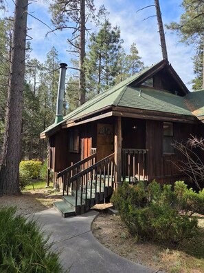 A rustic cabin with a green roof, a chimney, and a wooden porch with a staircase leading up to the entrance. - Cozy Studio in Idyllwild (Idyllwild-Pine Cove)