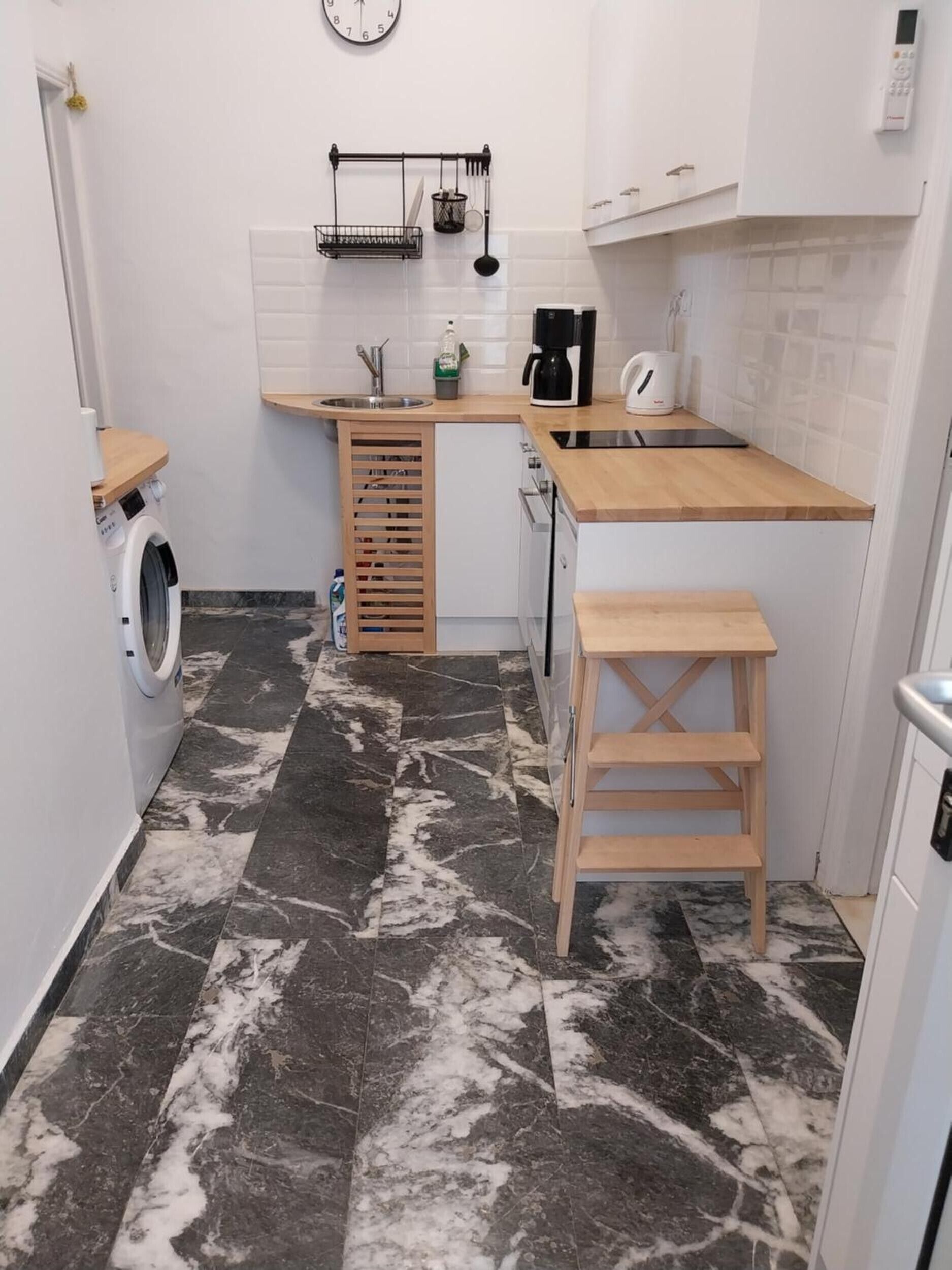 A compact kitchen with a washing machine, a sink, a coffee maker, and a wooden stool.