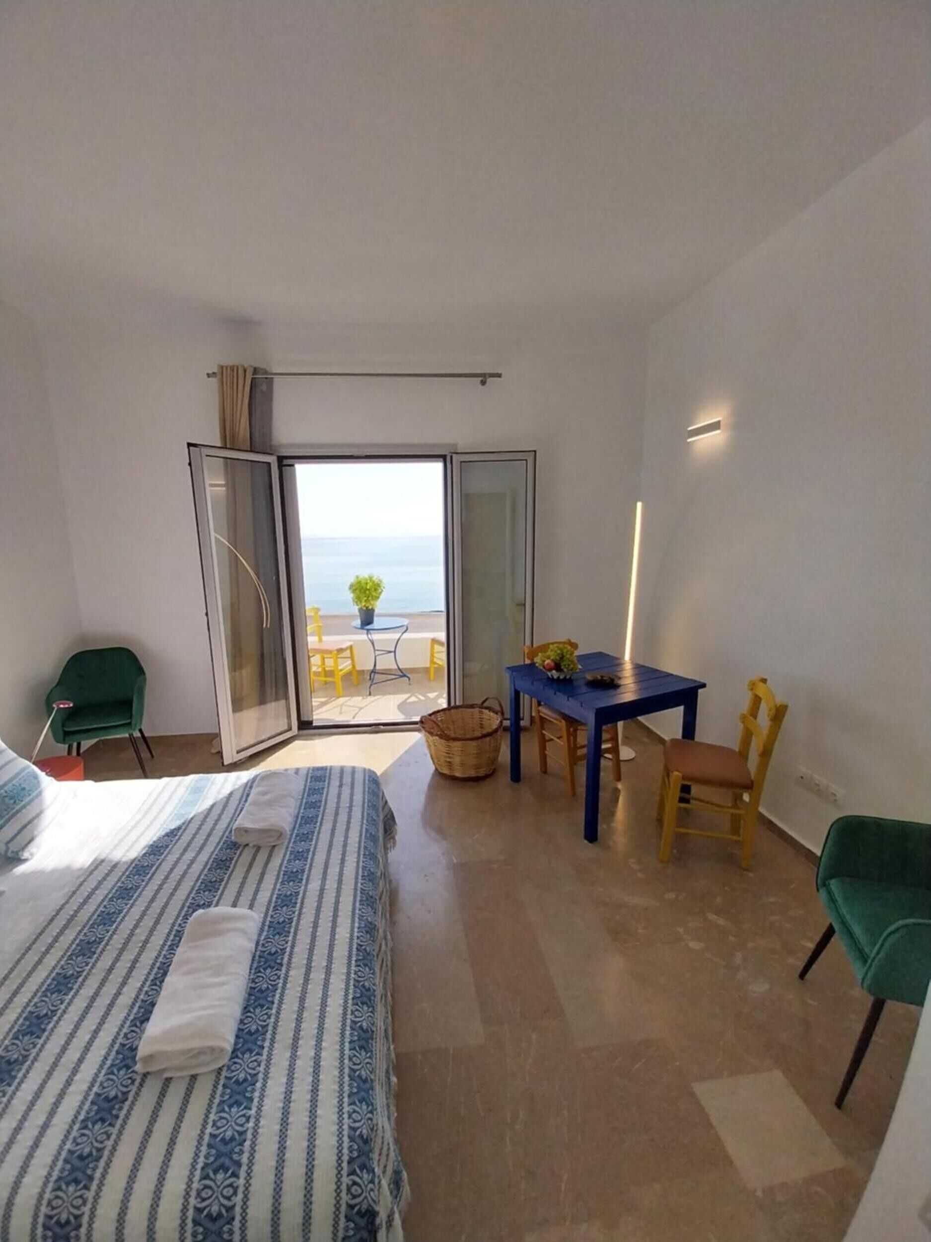 Habitació d'hotel amb llit, taula, cadires, taula petita amb una planta i vista al mar per una porta corredissa.