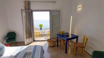 Habitació d'hotel amb llit, taula, cadires, taula petita amb una planta i vista al mar per una porta corredissa.