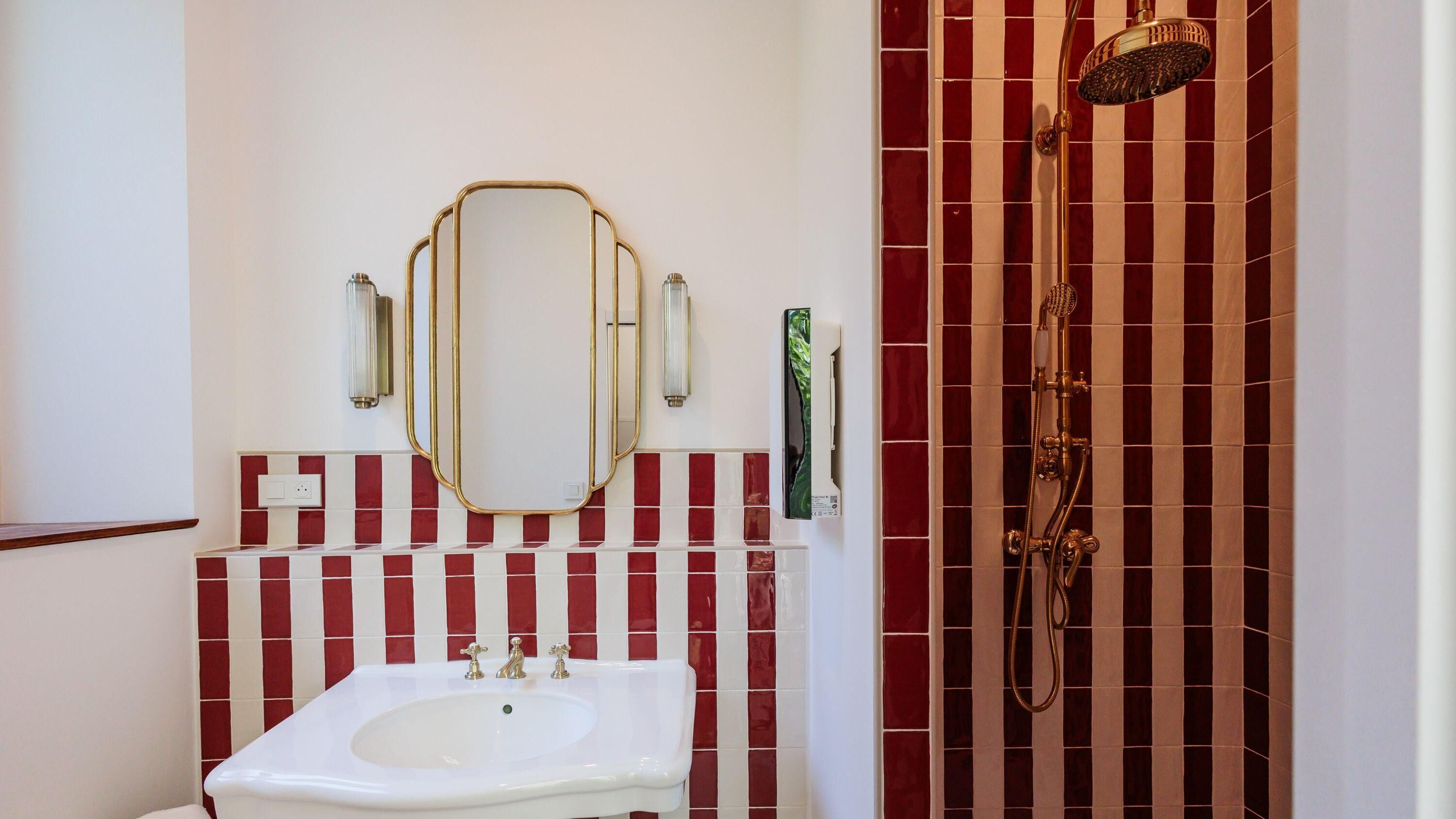 Un baño con un lavamanos blanco, un espejo con marco dorado, azulejos a rayas rojas y blancas, una ducha con cabezal dorado y un dispensador de jabón verde.