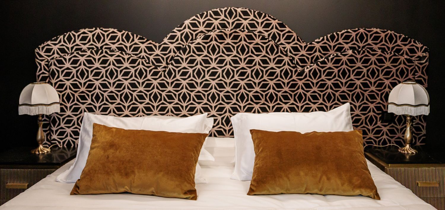 Habitación de hotel con cabecera estampada, dos almohadas blancas, dos almohadas marrones y dos lámparas de noche.