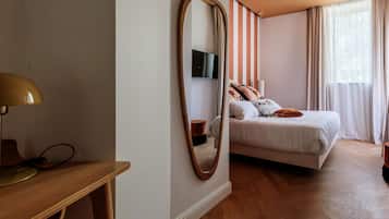 A bedroom with a bed, a desk with a lamp, a mirror, and a TV on the wall.