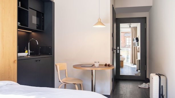 A hotel room with a bed, a small table with a chair, a microwave, a sink, and a suitcase. - STAYERY Fürth Innenstadt (Fuerth)