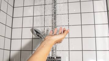 A hand reaching for a showerhead in a tiled bathroom with a mirror and two bottles on the wall.