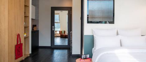 A modern hotel room with a bed, a small side table, a wardrobe, and a view into the adjacent living area.