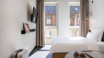 A hotel room with a bed, a desk, a laptop, a suitcase, a TV, and a view of a building through the window.