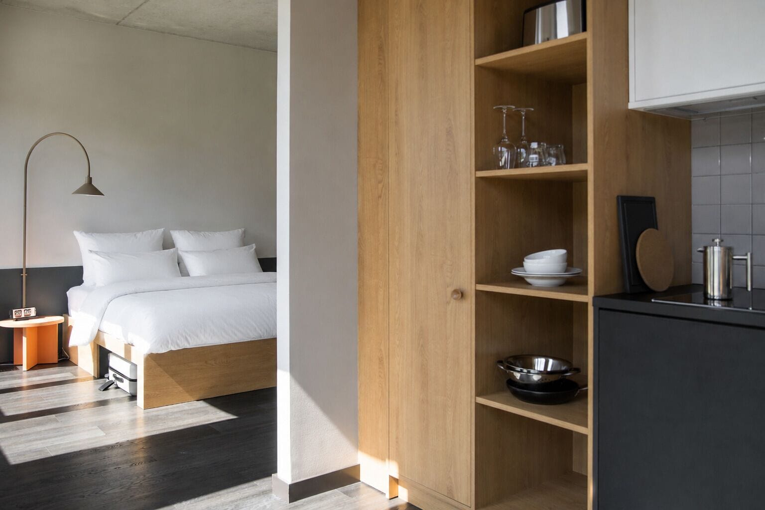 A modern hotel room with a bed, a wooden wardrobe, a kitchen area, and a bedside table with a lamp.