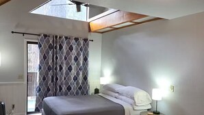 Une chambre à coucher comprenant un lit, des lampes de chevet, une petite table, un téléviseur, une fenêtre avec des rideaux et un puits de lumière.