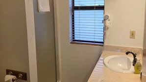 Une salle de bain avec une douche, une fenêtre avec des stores, un lavabo avec un distributeur de savon et une serviette accrochée à la porte.