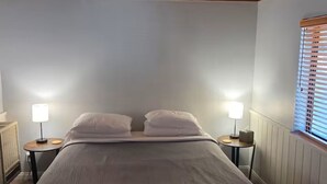 A bedroom with a wooden ceiling, a bed with white pillows, two bedside tables with lamps, and a window with blinds. - Comfortable 1-bedroom Suite (Idyllwild-Pine Cove)