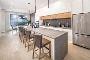Una cocina moderna con una isla central, taburetes de bar, electrodomésticos de acero inoxidable y un amplio ventanal con persianas.