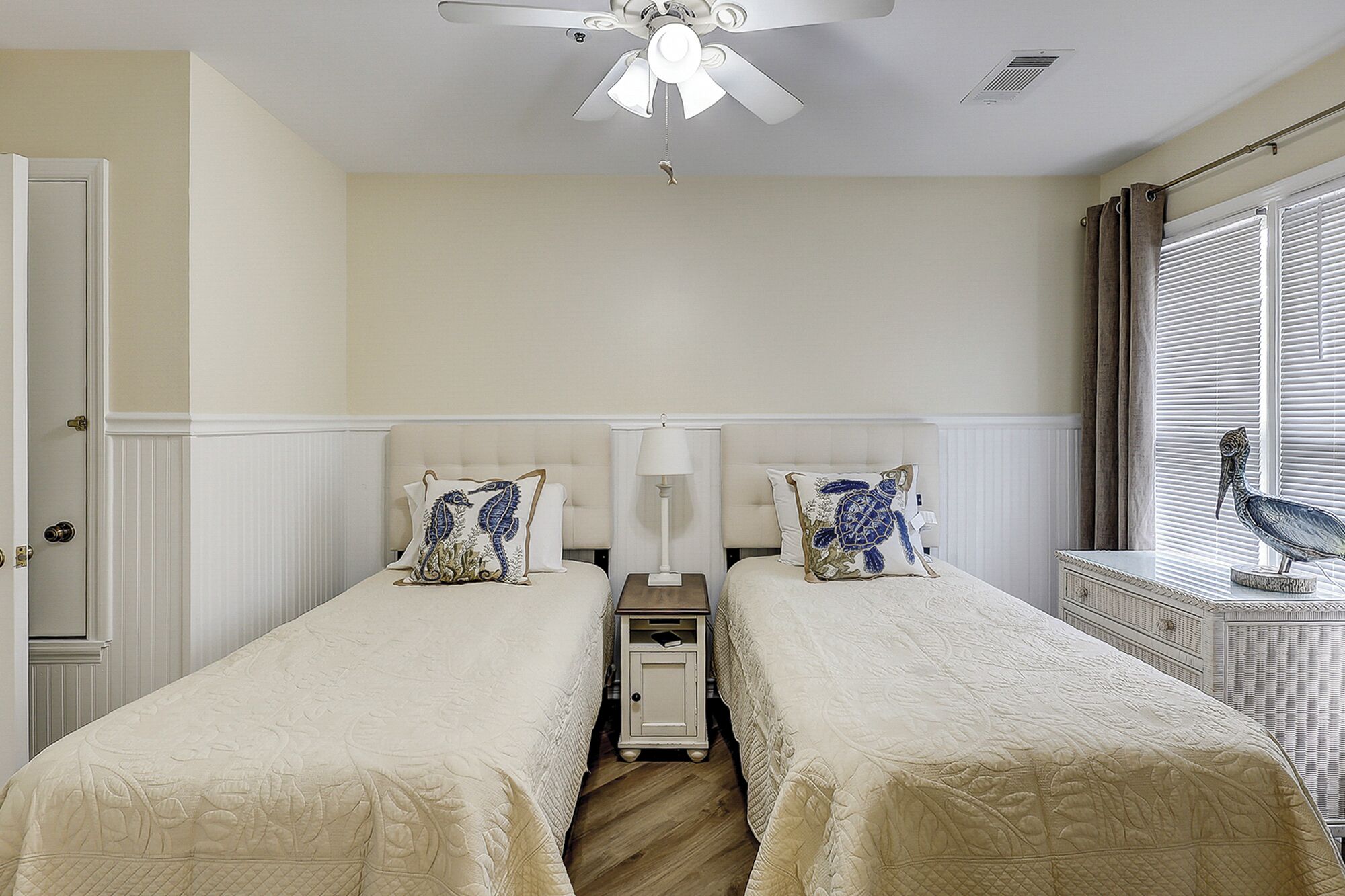 A room with two beds, a ceiling fan, a window with blinds, and a nightstand with a lamp.